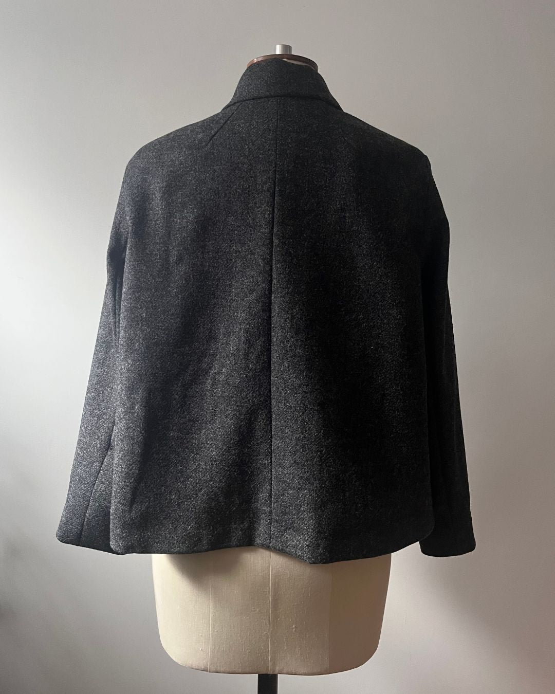 A dark gray textured wool cape jacket displayed on a cream-colored dress form mannequin, showing the back view against a neutral gray background.