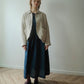 A woman stands in a minimalist room with a light gray wall and herringbone wood floor, wearing a beige linen button-up shirt jacket over a dark blue midi dress, paired with black Mary Jane shoes.