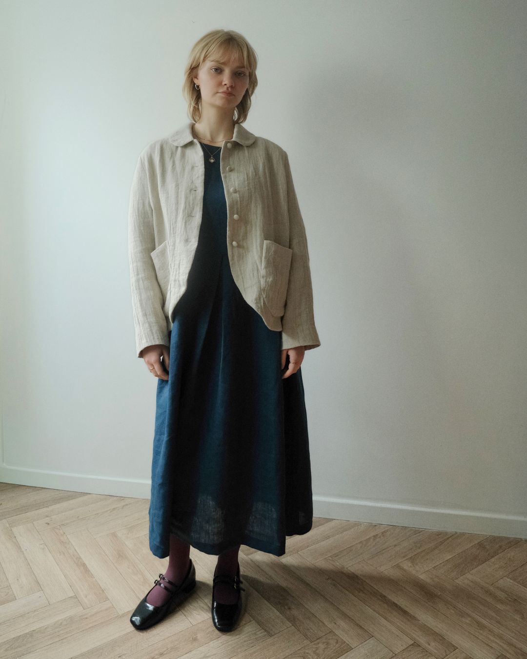 A woman stands in a minimalist room with a light gray wall and herringbone wood floor, wearing a beige linen button-up shirt jacket over a dark blue midi dress, paired with black Mary Jane shoes.