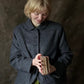 A woman with short blonde hair wearing a dark grey textured wool jacket over an olive green top, holding a small stack of books against a dark textured studio backdrop.