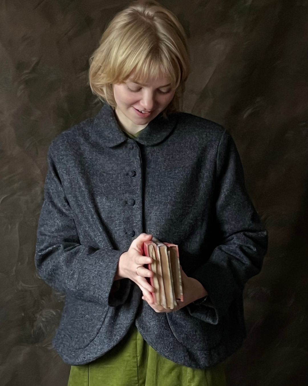 A woman with short blonde hair wearing a dark grey textured wool jacket over an olive green top, holding a small stack of books against a dark textured studio backdrop.
