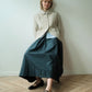 A woman stands against a plain off-white wall on a light wood herringbone floor, wearing a cream textured button-front cardigan over a light blue collared shirt and a dark teal wide-leg skirt, with black Mary Jane shoes.