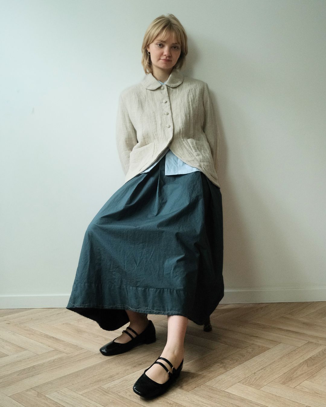 A woman stands against a plain off-white wall on a light wood herringbone floor, wearing a cream textured button-front cardigan over a light blue collared shirt and a dark teal wide-leg skirt, with black Mary Jane shoes.