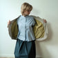 A woman stands in a room with a light-colored wall and wood floor, holding open a quilted jacket to show its olive green lining. She is wearing a light blue button-up shirt and a dark teal skirt.