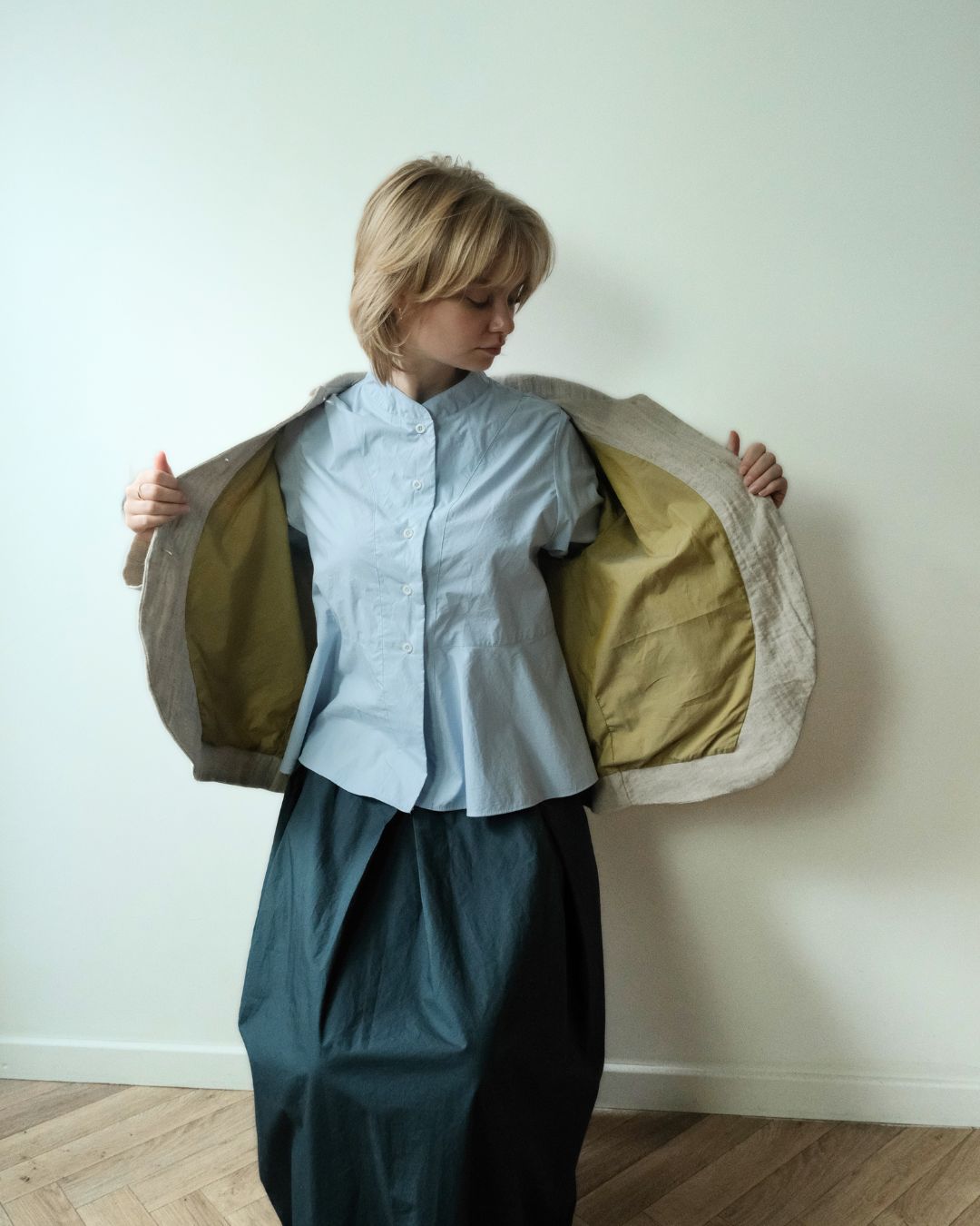 A woman stands in a room with a light-colored wall and wood floor, holding open a quilted jacket to show its olive green lining. She is wearing a light blue button-up shirt and a dark teal skirt.