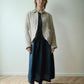 A woman stands in a minimalist room with a light wall and wood floor, wearing a beige linen button-up jacket over a dark navy midi dress with dark lace-up shoes.