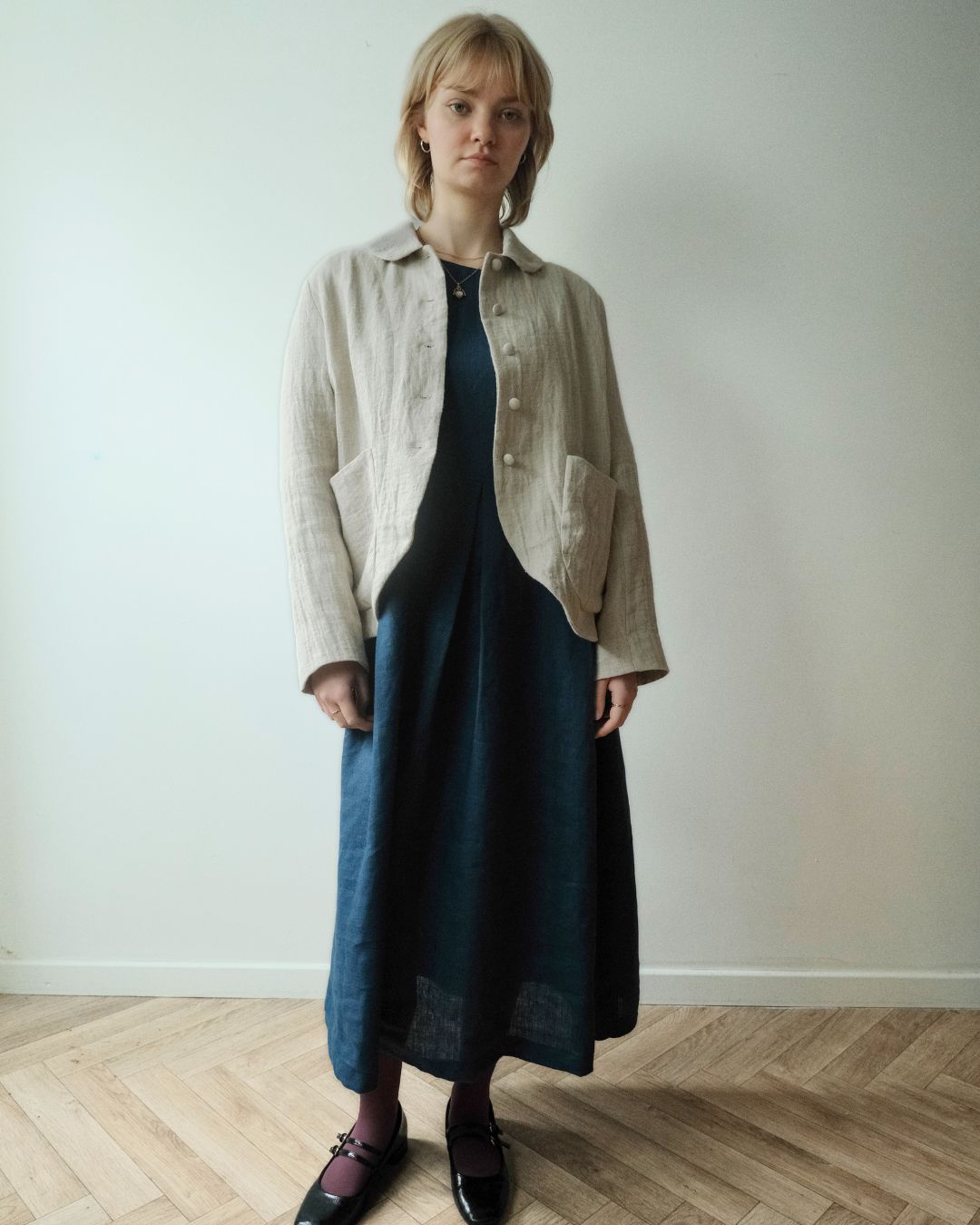 A woman stands in a minimalist room with a light wall and wood floor, wearing a beige linen button-up jacket over a dark navy midi dress with dark lace-up shoes.