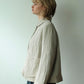 A woman in profile wearing a light beige linen jacket with a collar and front pockets, paired with dark blue skirt, standing against a plain off-white wall.