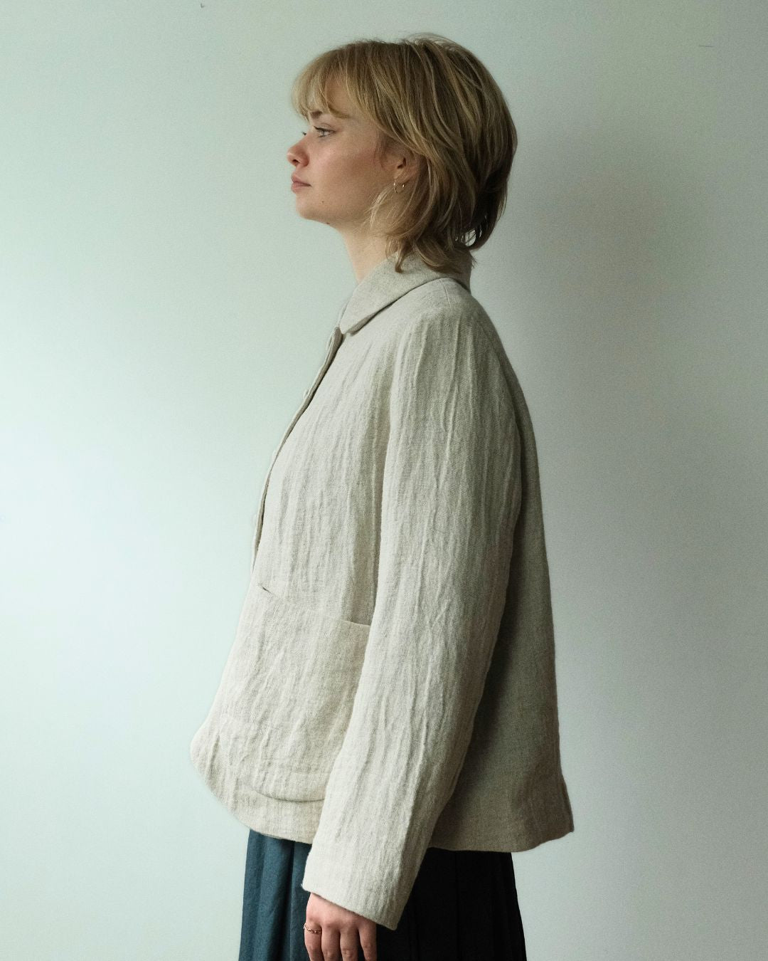 A woman in profile wearing a light beige linen jacket with a collar and front pockets, paired with dark blue skirt, standing against a plain off-white wall.