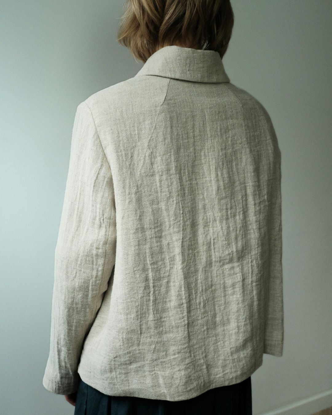 A person viewed from the back wearing a light beige linen-textured collared jacket or shirt against a plain light gray wall.