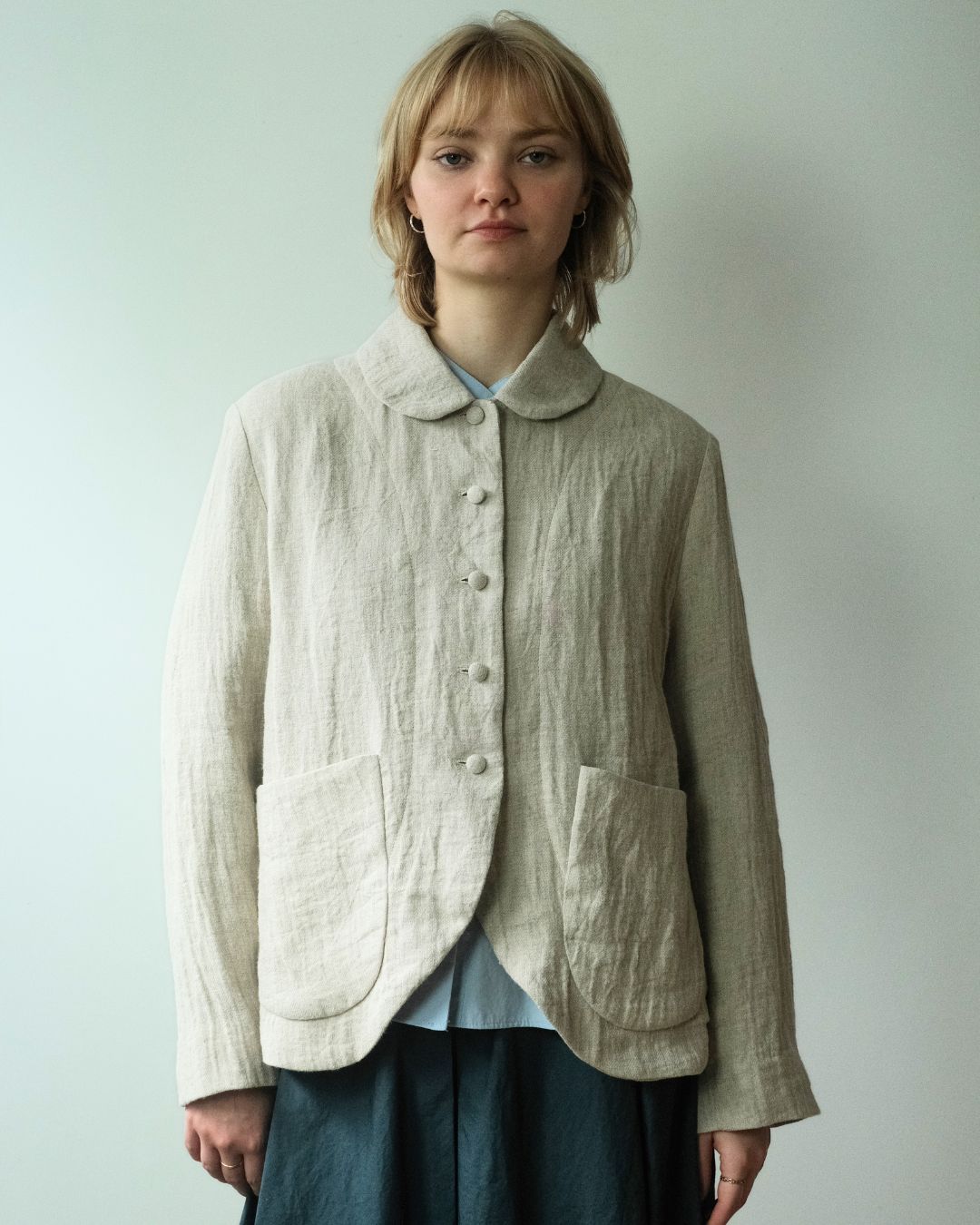 A woman wearing a light beige linen button-up jacket with a Peter Pan collar and large front patch pockets, layered over a light blue top and dark skirt, standing against a plain light gray wall.