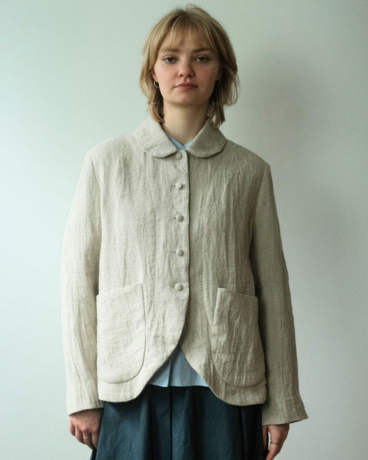 A woman wearing a light beige linen button-up jacket with a Peter Pan collar and large front patch pockets, layered over a light blue top and dark skirt, standing against a plain light gray wall.