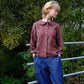 Person wearing a brown shirt and blue pants standing outdoors with greenery in the background