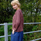 Woman wearing a maroon top and blue pants standing outdoors with greenery in the background