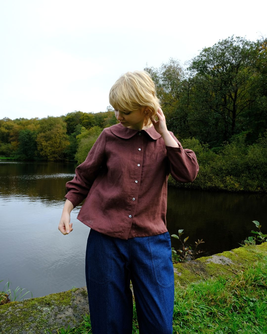 Person wearing a brown jacket and blue pants standing by a body of water with trees in the background