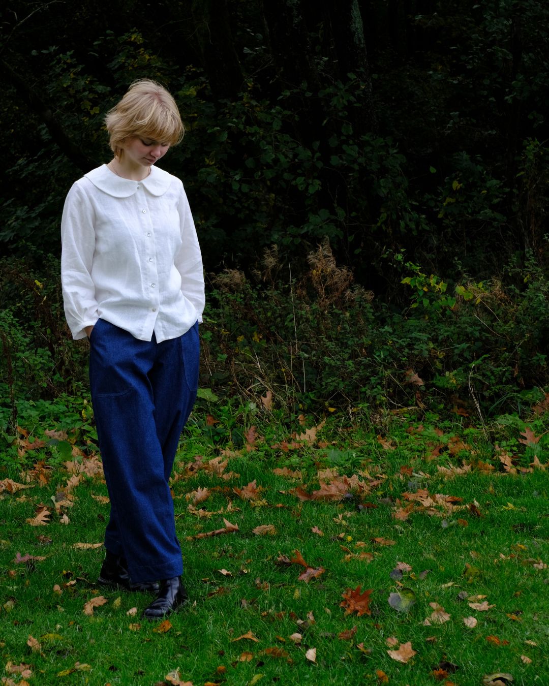 Person wearing a white blouse and blue pants standing in a grassy area with fallen leaves and greenery.