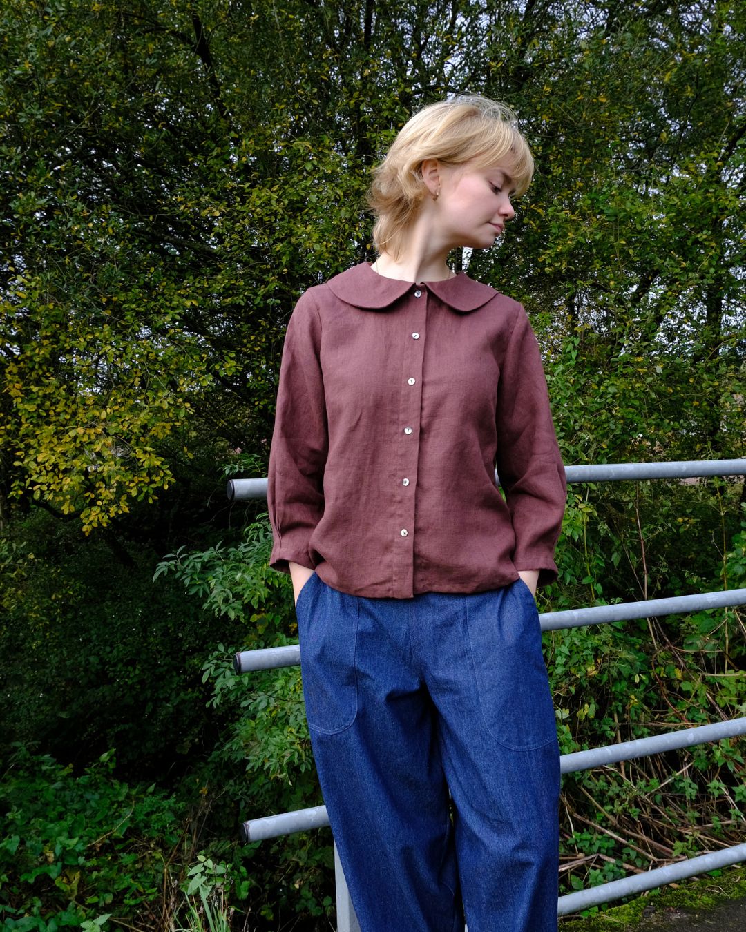 Person wearing a brown shirt and blue pants standing outdoors with greenery in the background