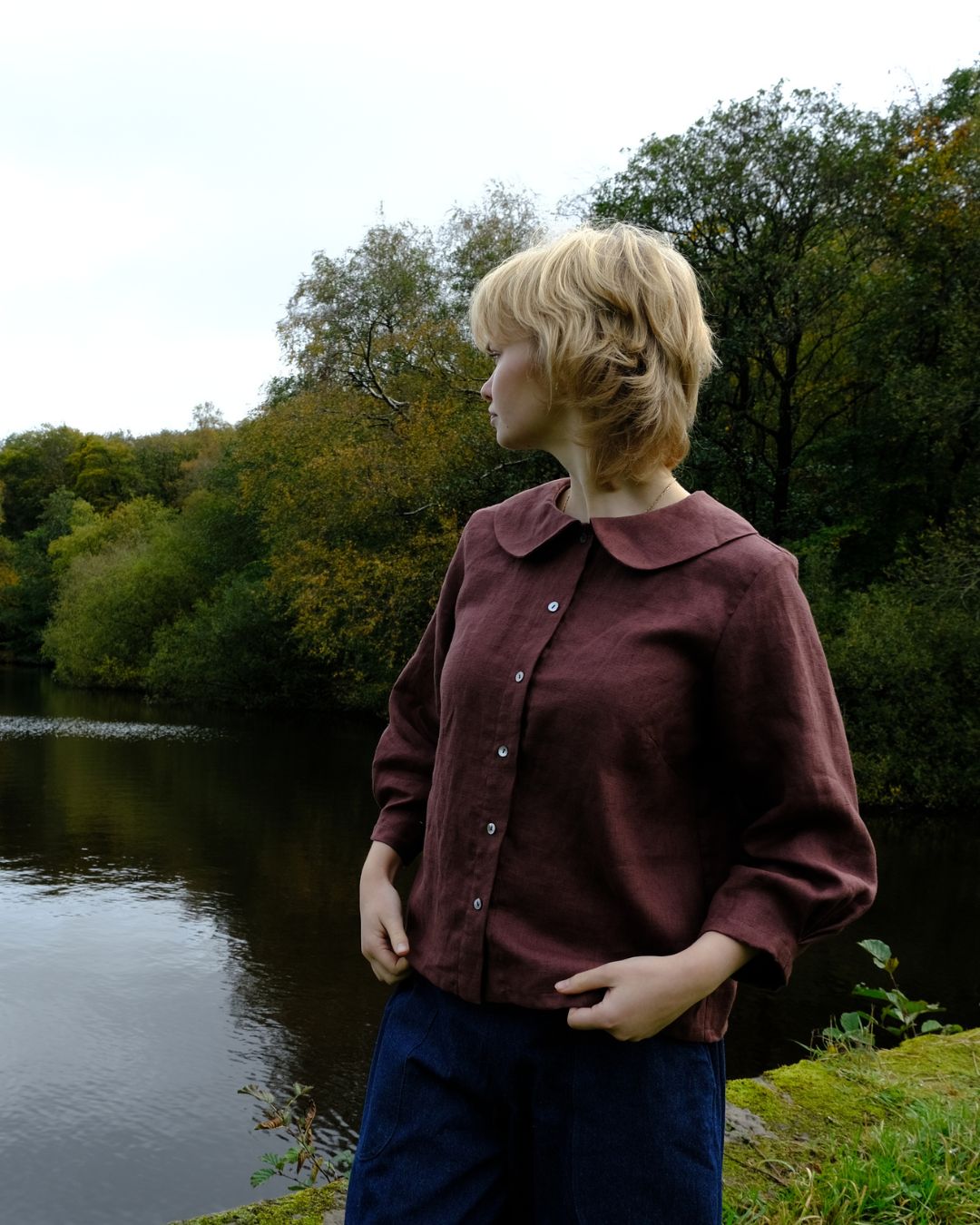 Person wearing a maroon shirt standing by a body of water with trees in the background