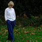 Person wearing a white blouse and blue pants standing in a grassy area with fallen leaves and greenery.