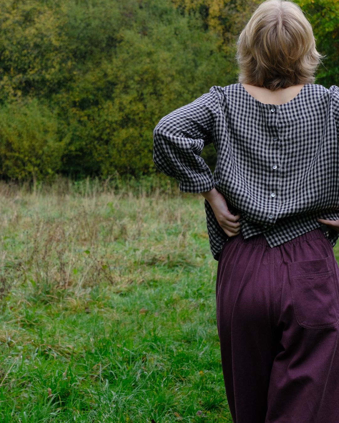 Person wearing a checkered shirt and purple pants standing in a grassy field with trees in the background