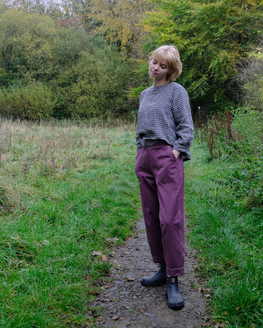 Person standing on a path in a natural setting wearing a checkered sweater and purple pants.