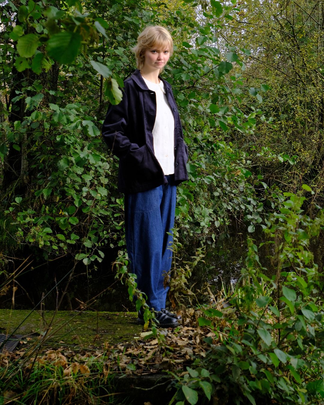 Person standing in a forested area wearing a black jacket and blue pants.