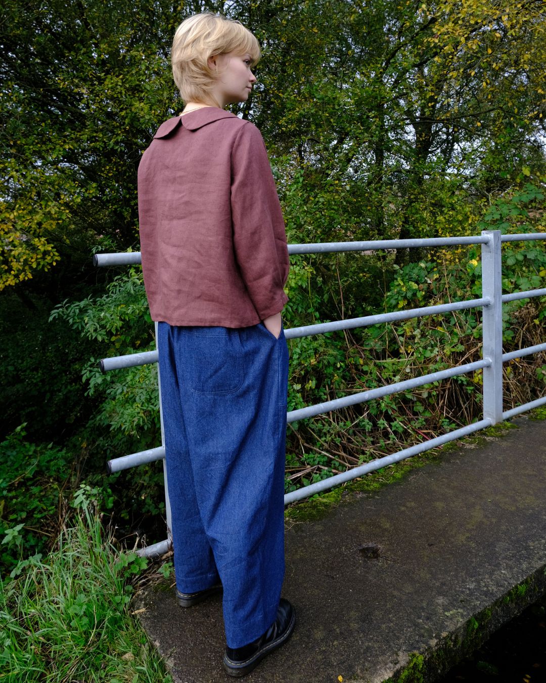 Person wearing a maroon jacket and blue pants standing near a metal railing with greenery in the background