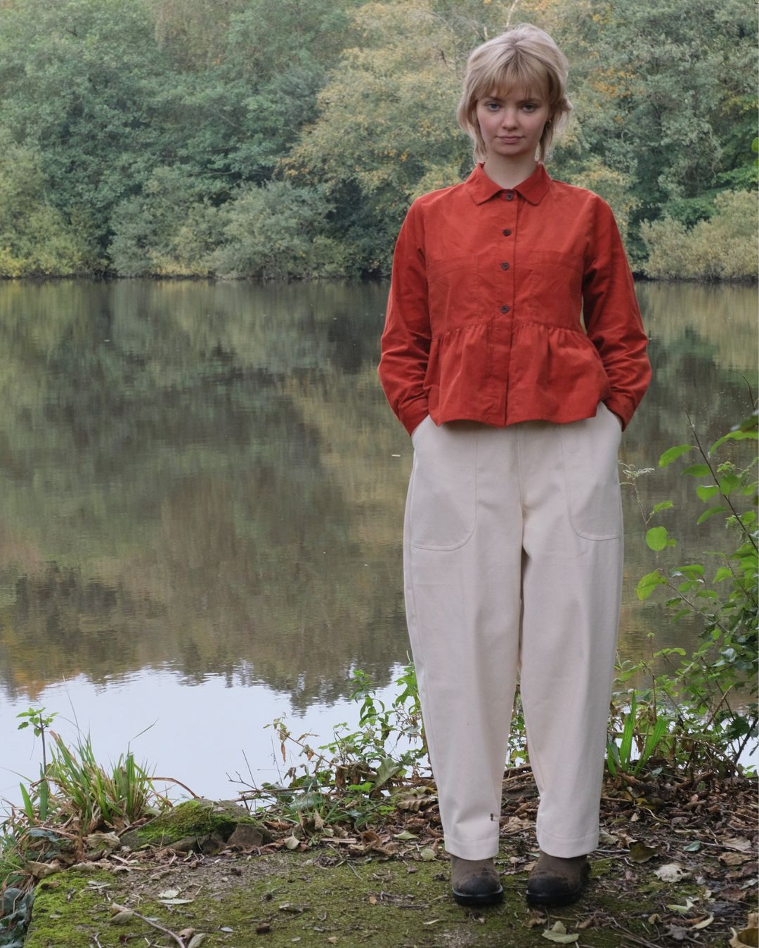 Person wearing a red jacket and white pants standing by a lake with trees in the background