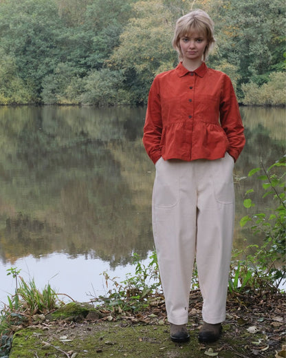 Person wearing a red jacket and white pants standing by a lake with trees in the background