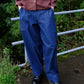 A person wearing loose blue denim pants, a brown button-up shirt, and black leather shoes, standing outdoors in front of a metal railing with green foliage.
