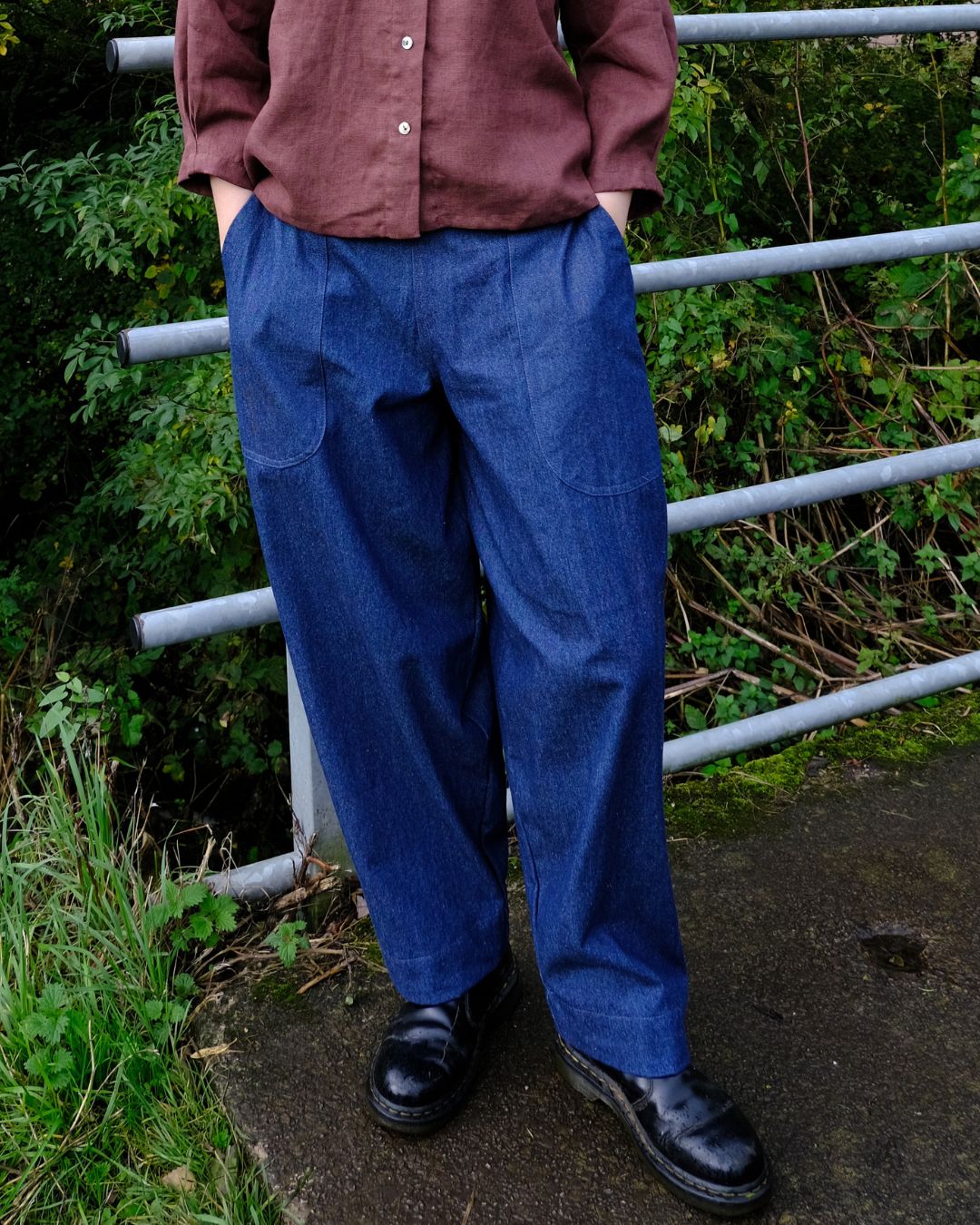 A person wearing loose blue denim pants, a brown button-up shirt, and black leather shoes, standing outdoors in front of a metal railing with green foliage.