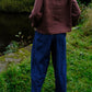 A person standing outdoors by a body of water, viewed from behind, wearing a loose brown top and dark blue wide-leg pants.