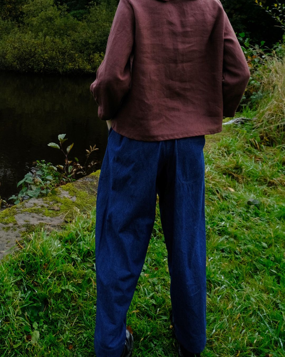 A person standing outdoors by a body of water, viewed from behind, wearing a loose brown top and dark blue wide-leg pants.