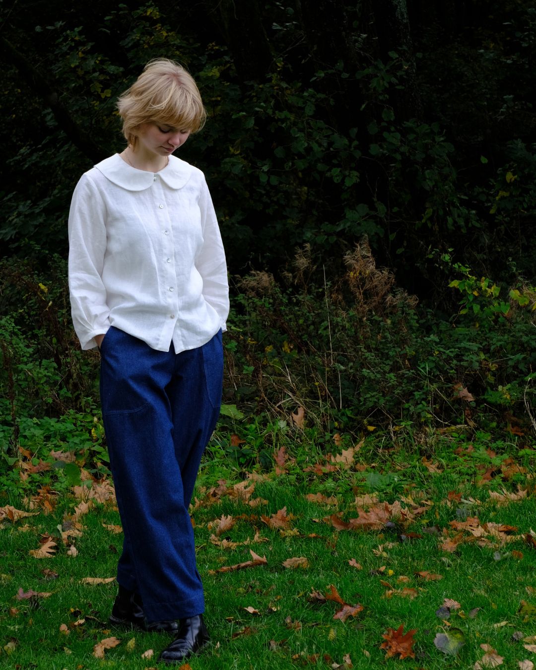 A woman standing outdoors on a grassy area with scattered autumn leaves, wearing a white long-sleeve button-up shirt with a wide collar and dark blue wide-leg trousers.
