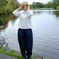A woman stands by a calm lake wearing a white long-sleeve button-up shirt and dark navy wide-leg trousers, with trees in the background.