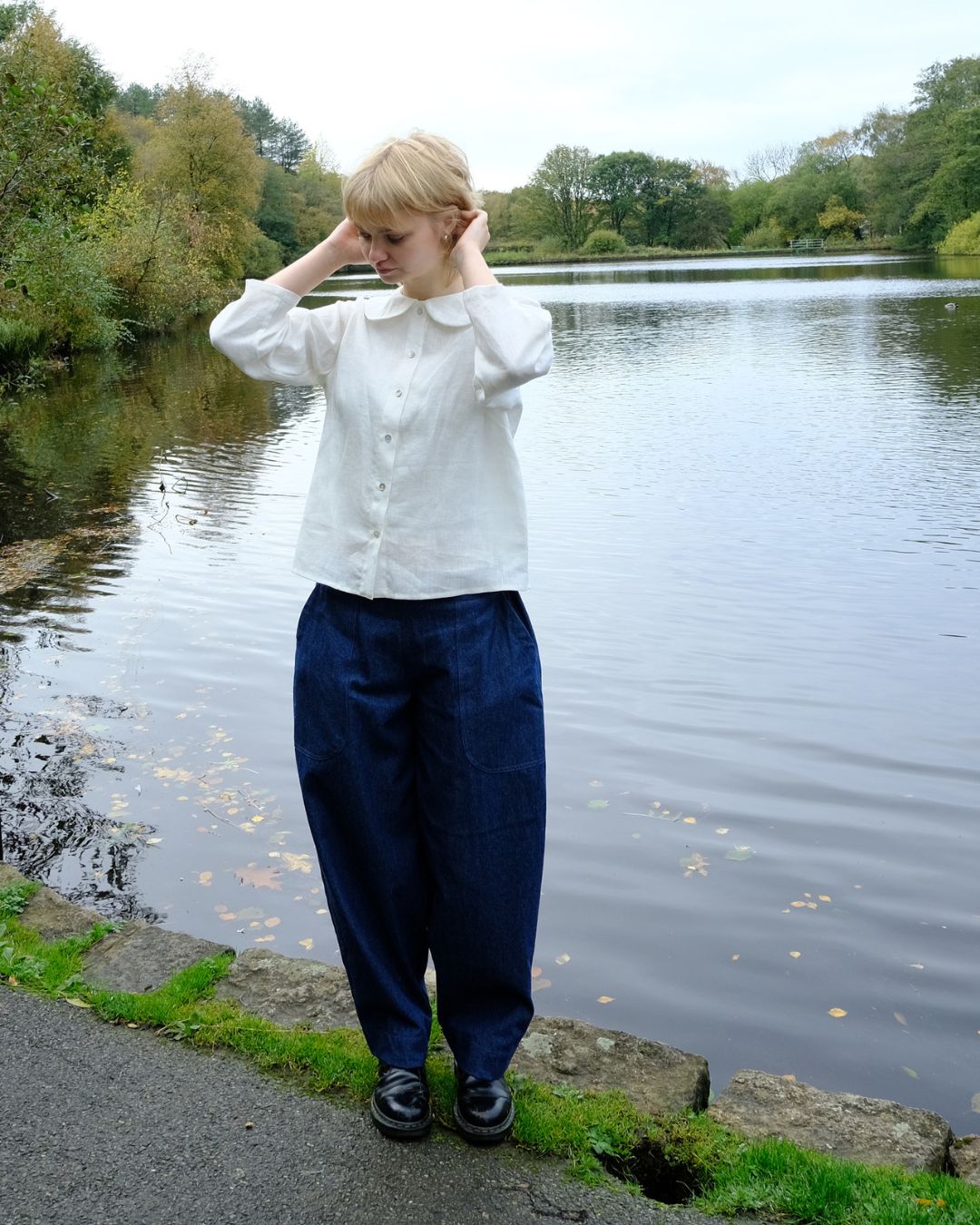 A woman stands by a calm lake wearing a white long-sleeve button-up shirt and dark navy wide-leg trousers, with trees in the background.