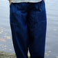 A person wearing dark indigo blue relaxed-fit pants with front patch pockets, paired with a white button-up shirt, standing outdoors near a body of water.