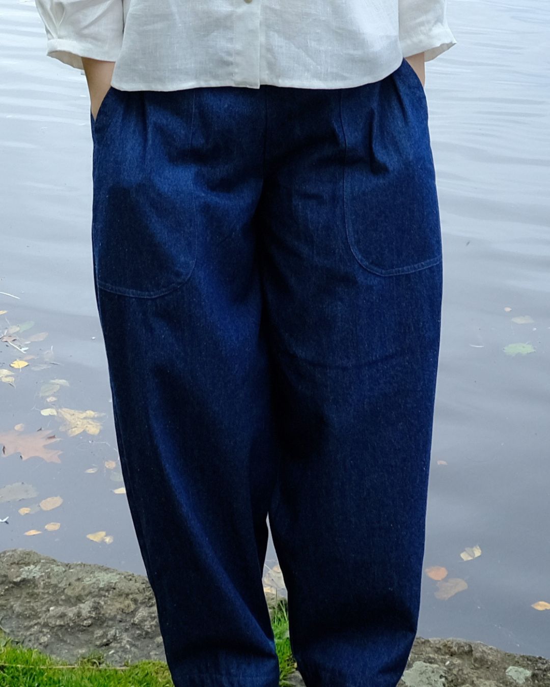 A person wearing dark indigo blue relaxed-fit pants with front patch pockets, paired with a white button-up shirt, standing outdoors near a body of water.