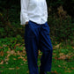 A person standing outdoors on grass with fallen leaves, wearing a white long-sleeve button-up shirt and loose dark blue trousers, with black shoes.