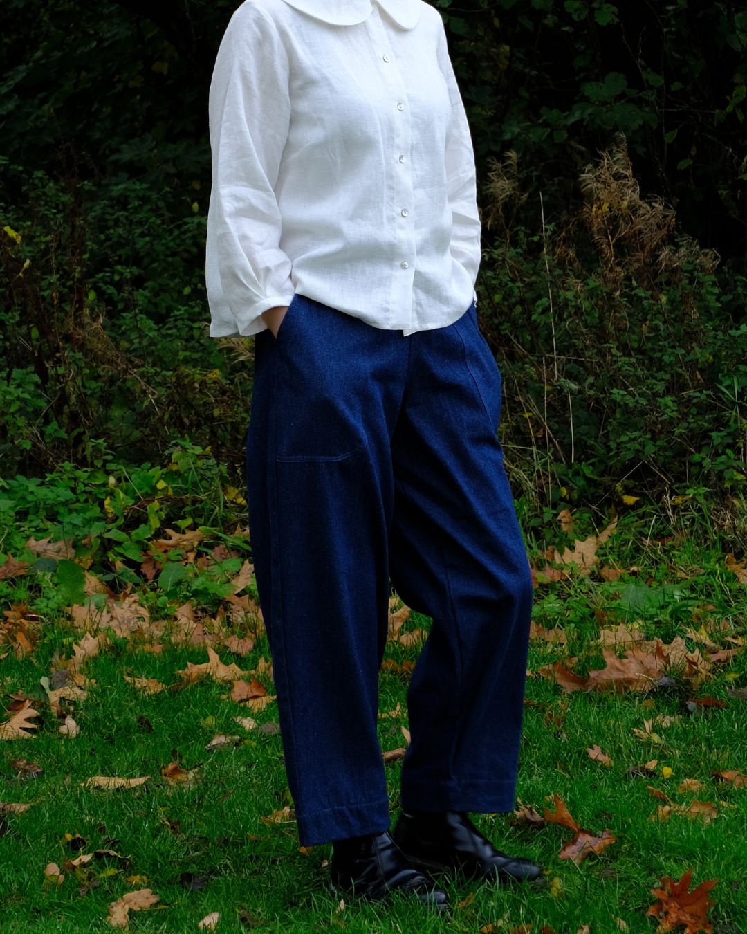 A person standing outdoors on grass with fallen leaves, wearing a white long-sleeve button-up shirt and loose dark blue trousers, with black shoes.