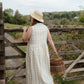 Woman in a white dress and straw hat standing near a wooden gate with a wicker basket, looking over a scenic landscape.