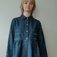 A model wearing a dark blue denim long-sleeve button-up shirt with white contrast stitching and decorative stitching on the chest pockets, paired with a black and white gingham skirt, set against a plain light gray wall.
