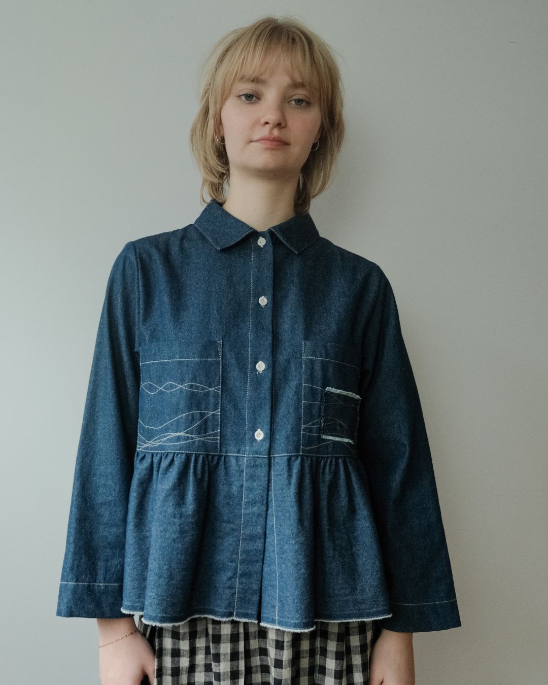 A model wearing a dark blue denim long-sleeve button-up shirt with white contrast stitching and decorative stitching on the chest pockets, paired with a black and white gingham skirt, set against a plain light gray wall.