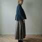 A woman stands in profile in a minimalist room with a light gray wall and wood herringbone floor, wearing a dark blue cropped ruffled jacket over a black and white gingham maxi skirt with black leather boots.