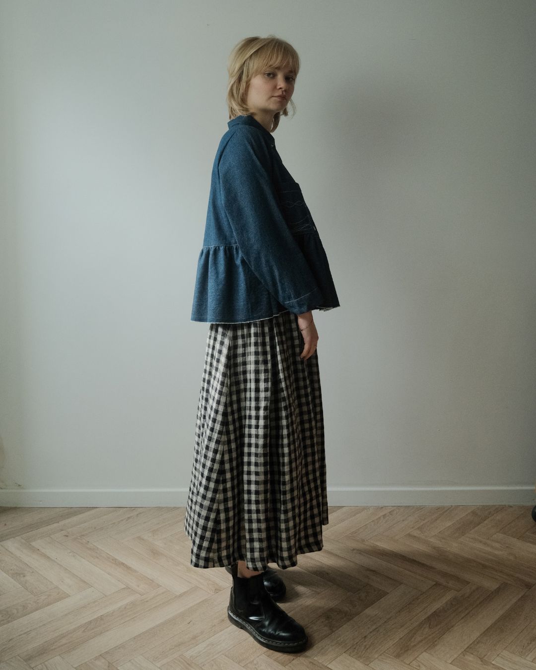 A woman stands in profile in a minimalist room with a light gray wall and wood herringbone floor, wearing a dark blue cropped ruffled jacket over a black and white gingham maxi skirt with black leather boots.