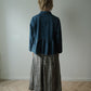 A person standing with their back to the camera, wearing a dark blue denim long-sleeve top with a ruffled hem and a black-and-white gingham midi skirt, in a minimalist room with a light gray wall and wooden floor.