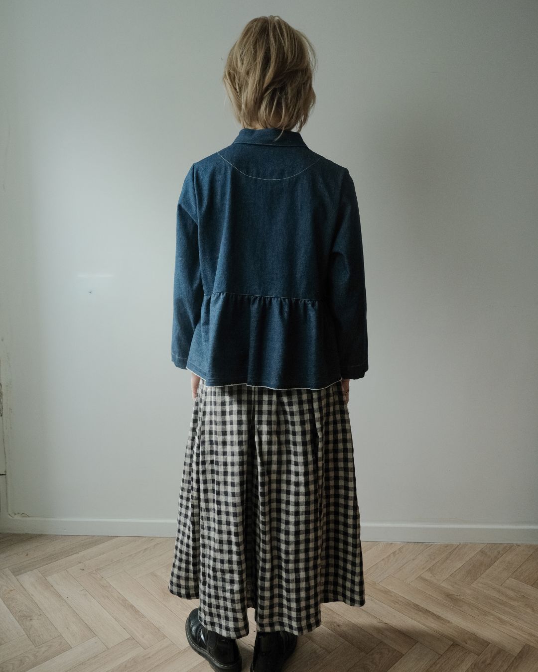 A person standing with their back to the camera, wearing a dark blue denim long-sleeve top with a ruffled hem and a black-and-white gingham midi skirt, in a minimalist room with a light gray wall and wooden floor.