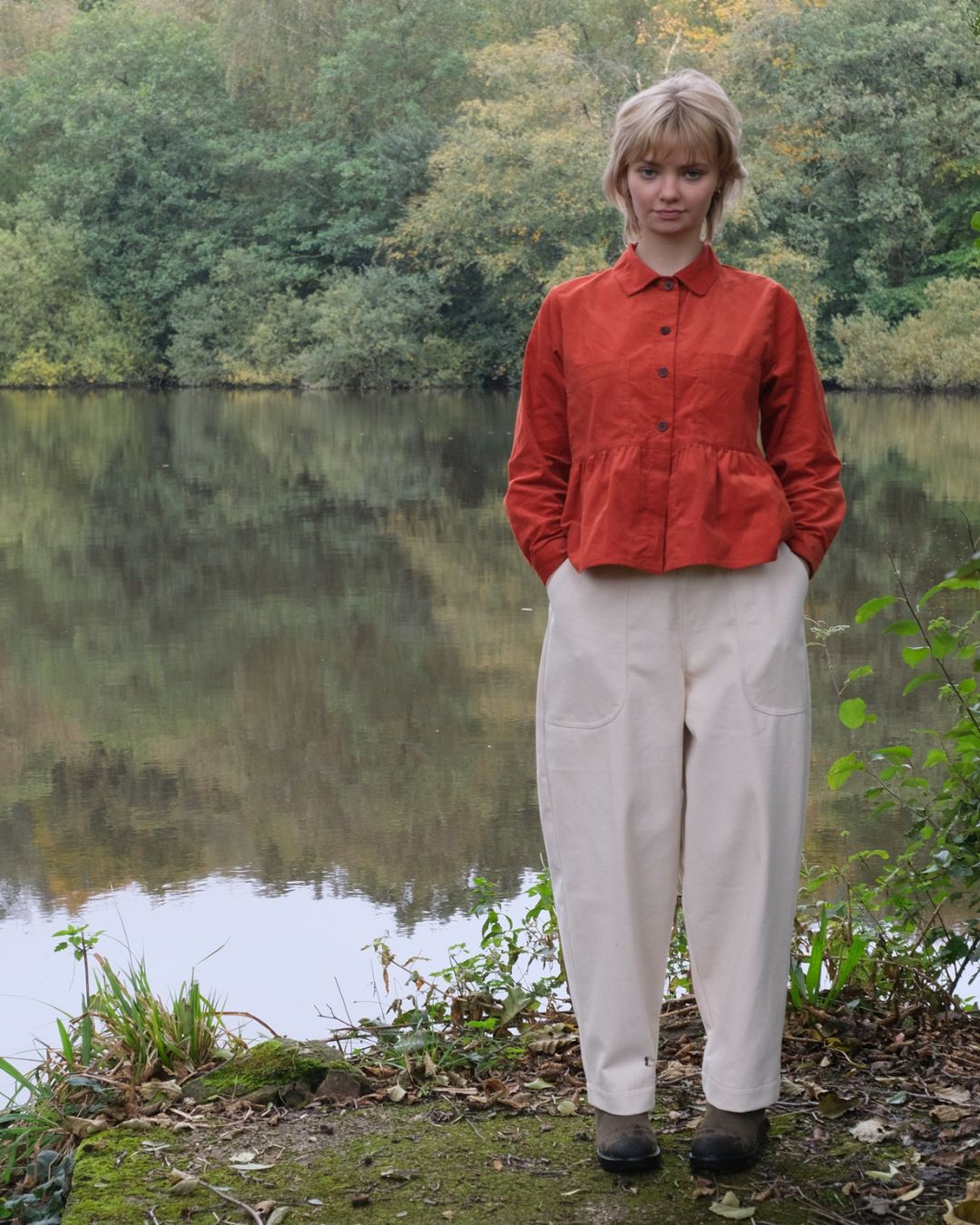 Person wearing a red jacket and white pants standing by a lake with trees in the background