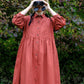 Person wearing a long rust-colored dress standing in front of green foliage.