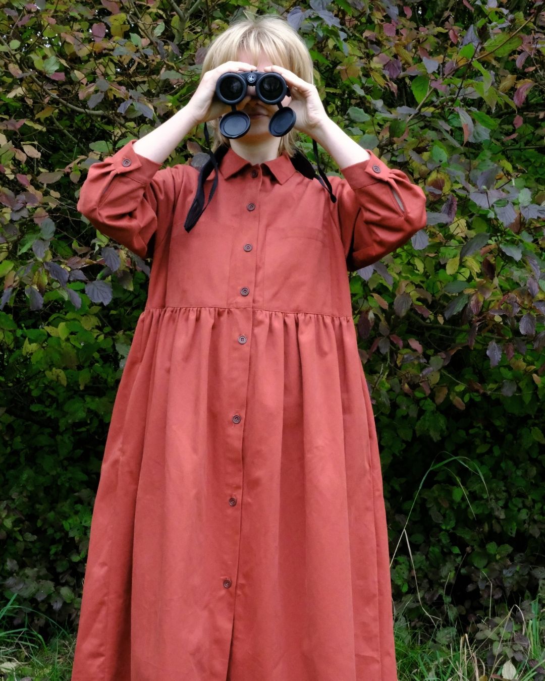 Person wearing a long rust-colored dress standing in front of green foliage.